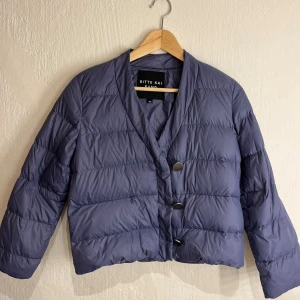 Blå jacka  - Säljer en blå pufferjacka från Bitte Kai Rand i storlek XS. Jackan har en croppad passform, v-ringad hals och tre stora knappar framtill. Perfekt för dig som gillar stilren design och vill ha en lätt jacka med cool look.