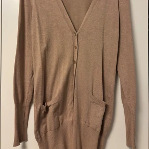 Beige kofta med v-ringning från Teenzies - Beiga kofta med djup v-ringning och knappar framtill. Går längre ner än ärmarna så är väldigt skön att ha om man vill täcka sig lite mer. Står inte storlek men skulle säga att den passar S-M💕