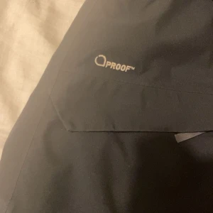 Svart Haglöfs vindjacka med huva - Svart vindjacka från Haglöfs med huva och hel dragkedja framtill. Jackan har reflekterande detaljer på ärmarna och är tillverkad i slitstarkt syntetmaterial. Perfekt för blåsiga dagar och outdoor-aktiviteter. Passar också S och xs