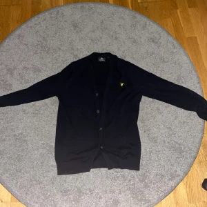 Mörkblå kofta från Lyle & Scott - Mörkblå  kofta från Lyle & Scott med klassisk gul logga på bröstet. Koftan har v-ringad hals, långa ärmar och knappar framtill. Perfekt för lager-på-lager och enkel att matcha med jeans eller chinos.