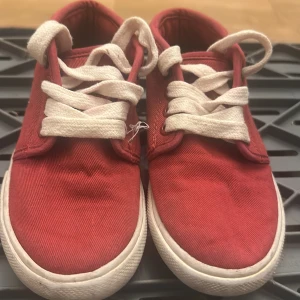 Röda sneakers från Lacoste - Snygga röda sneakers från Lacoste med vit sula och vita snören. Skorna har klassisk rund tå och är tillverkade i bomullstyg. På sidan finns den ikoniska gröna krokodilen broderad och Lacoste-loggan syns baktill. Perfekta för en avslappnad och sportig stil.