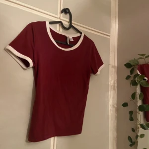 Vinröd t-shirt från H&M Divided XS - Säljer en vinröd t-shirt från H&M Divided i storlek XS. T-shirten har vita detaljer vid halsringning och vid ärmarna vilket ger en fin detalj på t-shirten. Perfekt topp i mjukt bomullsmaterial. Passar till jeans, mjukisbyxor eller annat!