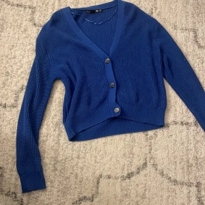 Blå kofta med v-ringning och knappar - Snygg blå kofta från laget157 med v-ringad hals och tre knappar framtill. Tröjan är stickad med ribbade muddar och har en croppad passform som ger en modern look. Perfekt att slänga över en t-shirt eller linne för extra stil.