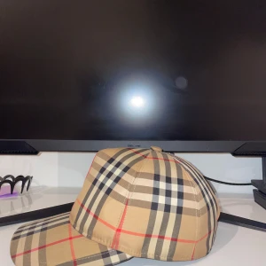 Burberry rutig keps i beige och svart - Säljer en klassisk Burberry keps med det ikoniska rutmönstret i beige, svart, vitt och röda linjer. Kepsen har böjd skärm, justerbar baksida och en patch med 'Burberry London England' bak. Perfekt accessoar för en trendig streetstyle-look.