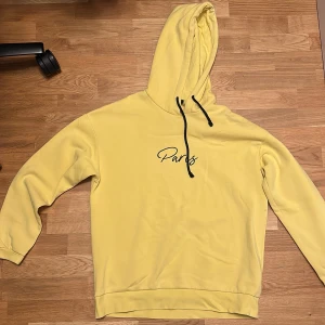 Gul hoodie Jack & Jones Paris XL - Snygg gul hoodie från Jack & Jones i storlek XL med svart snörning och trycket 'Paris' på bröstet. Mjuk insida och klassisk huva gör den perfekt för chill dagar. Passformen är normal och materialet är bomull med inslag av polyester.