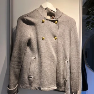 Grå skepparkavaj från Holly & Whyte - Snygg grå skepparkavaj med guldknappar från Holly & Whyte. Jackan har en stilren ståkrage, två fickor fram och är helfodrad. Materialet är mjukt och känns som ullmix. Perfekt för dig som gillar klassisk stil med en modern twist. Aldrig kommit till användning 