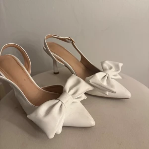 Balklackar - Snygga vita pumps med spetsig tå och hög klack. Skorna har en stor rosett framtill och en öppen häl med justerbart spänne. Tillverkade i skinnliknande material med en elegant och feminin look. Perfekta för dig som vill sticka ut med en chic detalj.