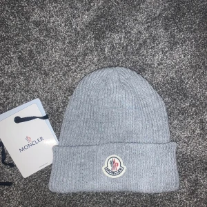 Grå Moncler mössa - Snygg grå ribbstickad mössa från Moncler med uppvikt kant och klassisk Moncler-logga framtill. Perfekt för kalla dagar och ger en clean look. Materialet är mjukt och stickat, vilket gör den både bekväm och stilren🤩