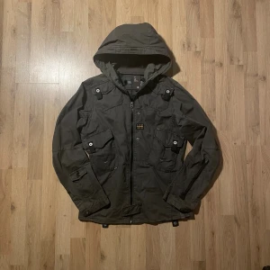 Mörkgrön fieldjacka från G-Star RAW - Snygg mörkgrön fieldjacka från G-Star RAW med huva, flera fickor med lock och dragkedja framtill. Jackan är tillverkad i bomull och har coola detaljer som knappar och patch på bröstet. Perfekt för dig som gillar streetwear och funktionella plagg.  Bilder på måtten finns tillgängliga. Fråga gärna! Snabb leverans!