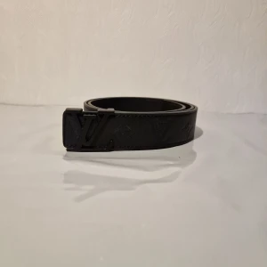 Svart Louis Vuitton skärp i skinn 110 cm - Snyggt svart bälte från Louis Vuitton med klassisk LV-logga som spänne och diskret mönster längs hela remmen. Bältet är tillverkat i skinn och har en stilren, modern look som passar till jeans eller kostym. Perfekt för dig som gillar exklusiva accessoarer.