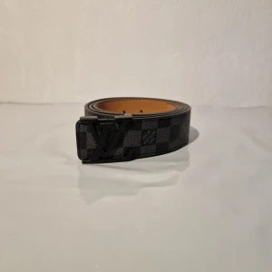 Svart rutig Louis Vuitton bälte 120 cm - Snyggt svart bälte från Louis Vuitton med klassiskt rutigt mönster och en stor LV-logga i metall som spänne. Bältet har en stilren design och insidan är beige. Perfekt för att lyfta din outfit med en lyxig touch.