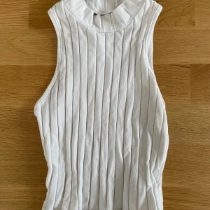 Vit ribbad ärmlös polotopp - Säljer en vit ärmlös polotopp med ribbad struktur och hög krage. Toppen har en clean och stilren look med vertikala ränder i tyget, perfekt för att skapa en fräsch och modern outfit.