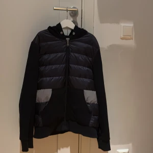 Moncler cardigan - Svart hoodie från Moncler med quiltad front och två fickor med grå detaljer. Tröjan har huva, långärmad design och dragkedja framtill. Moncler-logga på ärmen ger en exklusiv touch. Perfekt för dig som gillar stilrena och sportiga plagg. Denna cardigan är garanterat äkta och äkthetsbevis samt kvitto finns 