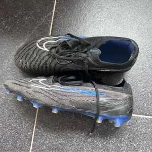 Nike Phantom  elite - Säljer ett par Nike Phantom Elite fotbollsskor i svart med blå och vita detaljer. Skorna har snörning, stickad ovandel och mönstrad struktur för bättre bollkontroll. Sulan är blå med fasta dobbar. Se bilder för defekter. Hör av dig vid fundering!😊🤩
