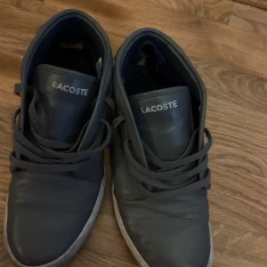 Gråa sneakers från Lacoste - Säljer ett par äkta gråa sneakers från Lacoste med klassisk design och snörning. Skorna har rund tå och är tillverkade i skinn, vilket ger en stilren och tidlös look. Perfekta för dig som gillar enkel och sportig stil.