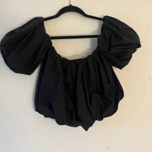 Svart blus med puffärmar - Säljer en svart blus med riktigt stora puffärmar och elastisk halsringning. Toppen har en loose passform och är tillverkad i ett lätt bomullstyg. Perfekt för dig som vill sticka ut med en cool och trendig siluett.