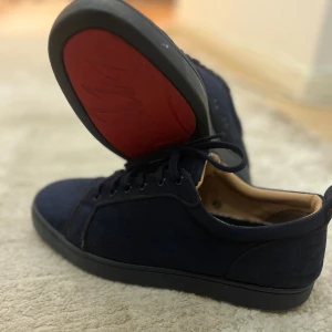 Mörkblå sneakers från Louboutin - Säljer ett par mörkblå sneakers från Christian Louboutin med klassisk röd sula och snygg mocka-look. Skorna har rund tå, snörning och diskreta metalldetaljer. Insidan är ljusbeige och sulan har den ikoniska Louboutin-signaturen.