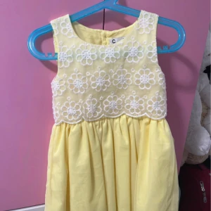 Gul ärmlös klänning med spets - Söt gul klänning med ärmlös design och rund hals. Överdelen har vit spets med blommigt mönster och kjolen är lätt rynkad för extra volym. Perfekt för sommarens alla tillfällen. Stängs med knapp bak.