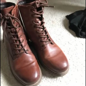 Bruna snörade boots i skinn Tamaris  - Snygga bruna boots med snörning och dragkedja på sidan för enkel påtagning. Tillverkade i slätt skinn med rund tå och platt sula. Perfekta för höst och vinter, med robust yttersula som ger bra grepp. Stilren design som passar till jeans och jacka.