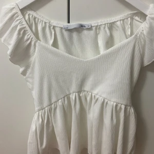 Vit offshoulder kortröja 157 - Superfin vit tröja från 157 med offshoulder och volangdetaljer. Tröjan har en ribbad struktur och flowy känsla. Perfekt för varma dagar och har en söt, romantisk vibe.