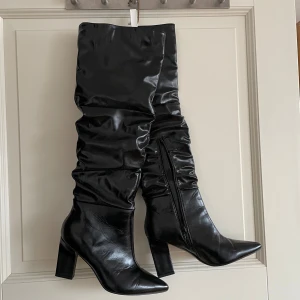 Svarta höga stövlar med klack - Snygga svarta stövlar i glansigt skinnliknande material med hög klack och spetsig tå. Stövlarna har dragkedja på insidan och en rynkad, slouchy design som går upp till knäet. Perfekta för att lyfta vilken outfit som helst.