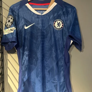 Chelsea Palmer 10 matchtröja Nike - Chelsea FC matchtröja med Palmer och nummer 10 på ryggen. Blå med subtilt mönster, vita detaljer och röd insida på kragen. Kortärmad, officiell UEFA Champions League-märke på ärmen. Tillverkad i lätt, ventilerande material med Dri-FIT-teknologi. Helt ny med tagg. En replika