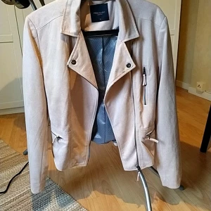 Ljus gammelrosa mockaliknande bikerjacka Freequent - Snygg ljus gammelrosa  bikerjacka från Freequent i mockaliknande material. Jackan har klassisk krage, asymmetrisk dragkedja och två fickor med dragkedjor framtill. Perfekt för dig som vill ha en stilren och trendig look.                                       Axelmått 44 cm längd 55 cm