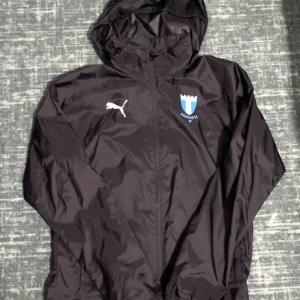Svart Malmö FF vindjacka från Puma - Svart vindjacka från Puma med Malmö FF-logga på bröstet. Jackan har huva, hel dragkedja framtill och två sidofickor. Tillverkad i lätt polyester, perfekt för dig som vill visa support för MFF. Klassisk sportig look med vit Puma-logga.