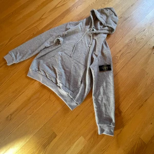 Grå hoodie från Stone Island - Snygg grå hoodie från Stone Island med klassisk logopatch på ärmen. Tröjan har huva med snörning, stor magficka och ribbade muddar. Perfekt för dig som gillar streetwear och vill ha en stilren look. Materialet är mjuk bomull som känns skönt mot huden.