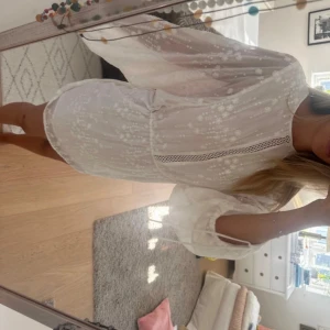 Vit kortklänning med volangärmar - Supersöt vit kortklänning med transparenta, voluminösa långärmar och diskret prickigt mönster. Klänningen har en snygg spetsdetalj framtill och knytband vid ärmsluten. 