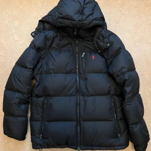 Svart dunjacka Polo Ralph Lauren M - Svart dunjacka från Polo Ralph Lauren med huva och dragkedja framtill. Jackan har en klassisk pufferlook, tre fickor med dragkedja och den ikoniska röda loggan broderad på bröstet. Perfekt för kalla dagar och riktigt snygg streetstil.