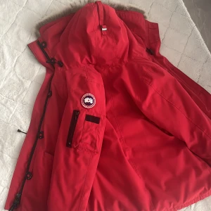 Röd Canada Goose äkta  dunjacka med pälskant - Säljer en röd Canada Goose dunjacka med huva och pälskant. Jackan har flera fickor, dragkedja och klassisk patch på ärmen. Insidan är svart och röd, och jackan är perfekt för kalla vinterdagar. Snygg och funktionell design som sticker ut.