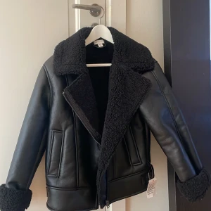 Svart teddyjacka från ASOS  - Säljer en svart teddyjacka från ASOS med bred krage och mjukt teddyfoder. Jackan har en cool oversized passform, dragkedja framtill och uppvikta ärmslut med teddy. Perfekt för dig som vill ha en trendig look.