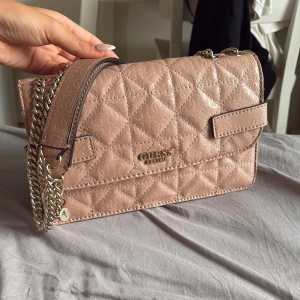 Beige quiltad axelväska från Guess - Snygg beige axelväska från Guess med quiltad design och subtilt logomönster. Väskan har guldfärgade kedjor, praktiskt fack med dragkedja på baksidan och flera innerfack. Perfekt storlek för det viktigaste och stilren look. NYPRIS 599kr