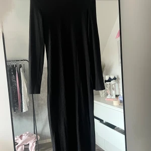 Svart ribbad långklänning - Säljer en svart långklänning med ribbad struktur och lång ärm. Klänningen har en rak och enkel siluett som ger en stilren look. Perfekt för dig som gillar minimalistisk stil och vill ha något som funkar till många tillfällen.