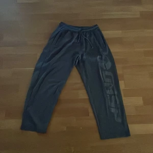 Mörkgrå mjukisbyxor med tryck - Säljer ett par mörkgrå mjukisbyxor med snörning i midjan och sidofickor. Ena benet har ett stort tryck längs sidan som ger en sportig vibe. Materialet är mjukt och skönt, perfekt för chill eller träning.