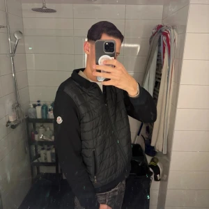 Svart cardigan från Moncler - Svart cardigan från Moncler med quiltad framsida och dragkedja. Cardiganen har två fickor med dragkedja och en klassisk Moncler-logga på ärmen. Materialet är bomull i ärmar och rygg, medan framsidan är dunvadderad för extra värme.