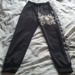 Svarta mjukisbyxor med lejontryck TA Fashion - Svarta mjukisbyxor från TA Fashion i storlek S med elastisk midja och ribbade muddar. Snyggt lejontryck i guld och vitt på ena benet samt dekorativt mönster längs sidorna. Mjukt material och en bakficka för extra stilpoäng.