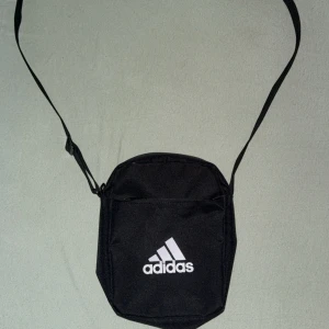 Svart axelväska från Adidas - Svart axelväska från Adidas med justerbar rem och klassisk vit logga på framsidan. Väskan har ett huvudfack med dragkedja och en mindre ficka framtill, perfekt för mobilen och småprylar. Tillverkad i slitstark polyester med stilren look. Även två olika fack i de stora facket vilket gör det lätt för dig.