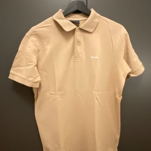 Beige pikétröja Peak Performance M - Snygg beige pikétröja från Peak Performance med klassisk krage och två knappar framtill. Broderad logga på bröstet. Tillverkad i bomull som känns skön mot huden. Perfekt för en clean och stilren look.
