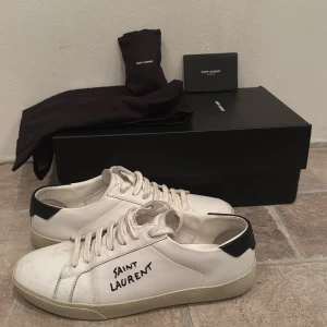 Saint Laurent vita sneakers i skinn - Snygga vita sneakers från Saint Laurent med svart häl och handskriven logga på sidan. Skorna har rund tå, platt sula och klassisk snörning. Tillverkade i skinn och levereras med originalkartong och dustbag. Perfekta för en clean och stilren look. Ingår även extra skosnören och brev. Det finns lite skador på skorna, hör av er vid funderingar.
