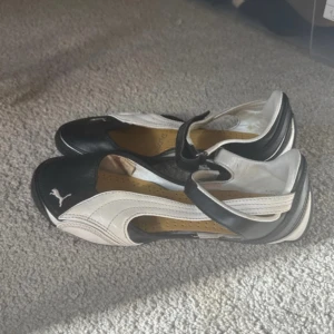 Svarta och vita ballerinaskor från Puma - Unika ballerinaskor från Puma i svart och vitt skinn med rund tå och coolt sportigt snitt. Skorna har en justerbar rem över vristen och klassisk Puma-logga framtill. Perfekta för dig som gillar en mix av sport och streetstyle.