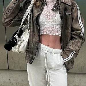 Brun skinnjacka från Collusion  - Säljer en brun skinnjacka från Collusion i bikerstil med vita ränder längs ärmarna och coola quiltade detaljer på axlar och armar. Jackan har ståkrage med tryckknapp och dragkedja framtill. Perfekt för dig som vill ha en edgy vibe.