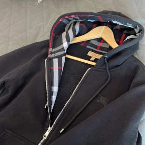 Svart hoodie från Burberry med rutig huva - Svart hoodie från Burberry i storlek L med klassisk rutig huva i grått, vitt, rött och svart. Tröjan har dragkedja, fickor framtill och broderad Burberry-logga på bröstet. Säljer då den blivit lite för liten för mig, bör passa dig perfekt om du är runt 180. Användes bara ett fåtal gånger innan jag blev för lång 🥲