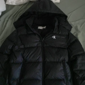 Svart pufferjacka Calvin Klein Jeans - Svart pufferjacka från Calvin Klein Jeans med huva och dragkedja. Jackan har tydlig CK-logga på bröstet och ett stort märke på ärmen. Fylld för extra värme och perfekt för kalla dagar. Klassisk och stilren design som funkar till allt.