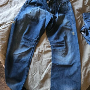 Blå jeans från Gasp med brodyr - Säljer ett par blå jeans från Gasp med coola sömdetaljer över knäna och broderad logga i gult på bakfickorna. Klassisk femficksmodell med bälteshällor och knappgylf. Perfekta för dig som gillar streetwear och vill sticka ut lite extra.