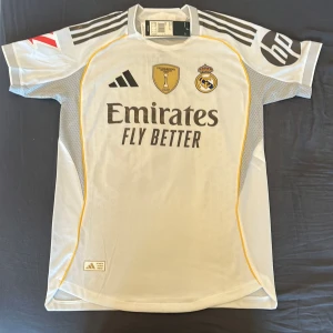 Real Madrid Mbappé 10 Adidas matchtröja - Säljer en officiell Real Madrid fotbollströja från Adidas med Mbappé och nummer 10 på ryggen. Tröjan är vit med svarta detaljer, guldiga inslag och gula linjer. Broderat klubbmärke, Champions-märke och sponsortryck. Tillverkad i ventilerande funktionsmaterial.