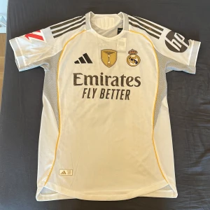 Real Madrid Mbappé 10 Adidas matchtröja - Säljer en officiell Real Madrid fotbollströja från Adidas med Mbappé och nummer 10 på ryggen. Tröjan är vit med svarta detaljer, gula inslag och klubbmärke broderat på bröstet. Tillverkad i ventilerande funktionsmaterial och har korta ärmar samt tryckta sponsorer.