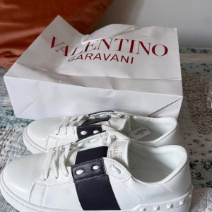 Valentino Garavani vita sneakers - Snygga vita sneakers från Valentino Garavani med bred svart rem över vristen och ikoniska nitar på hälen. Skorna har rund tå, platt sula och är tillverkade i skinn. Perfekta för dig som vill ha en clean men ändå edgy look.skorna är i storlek 42 och är helt nya