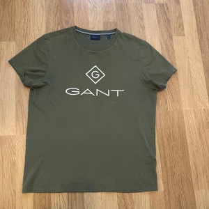 Olivgrön GANT t-shirt med logga - Snygg olivgrön t-shirt från GANT med vit logga tryckt på bröstet. Klassisk rund hals och korta ärmar. Tillverkad i mjuk bomull för en skön känsla. Perfekt för en avslappnad och stilren look.